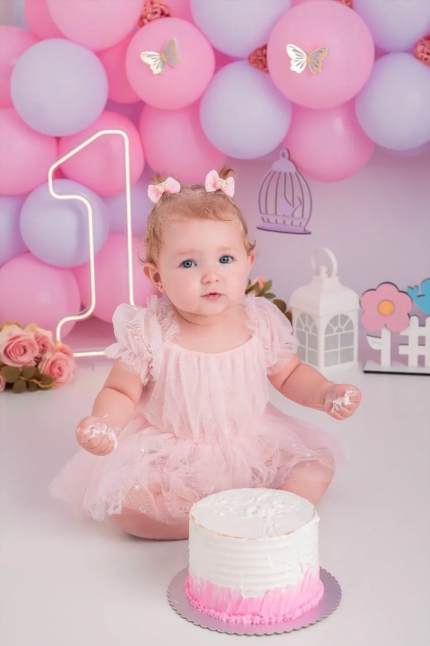 Bebê de vestido rosa de tule com as mãos sujas de chantilly diante do bolo, balões rosa e lilás ao fundo