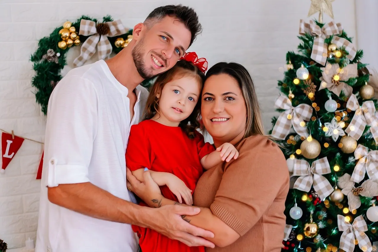 Família abraçada com a filha pequena de vestido vermelho ao lado da árvore de Natal