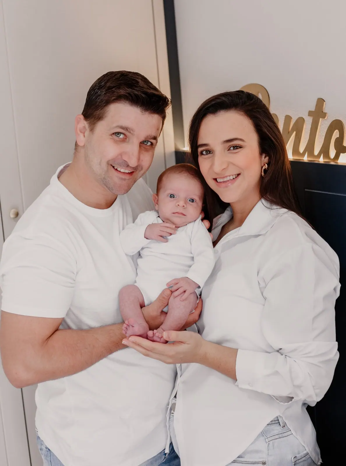 Pai e mãe de branco sorriem segurando o bebê recém-nascido