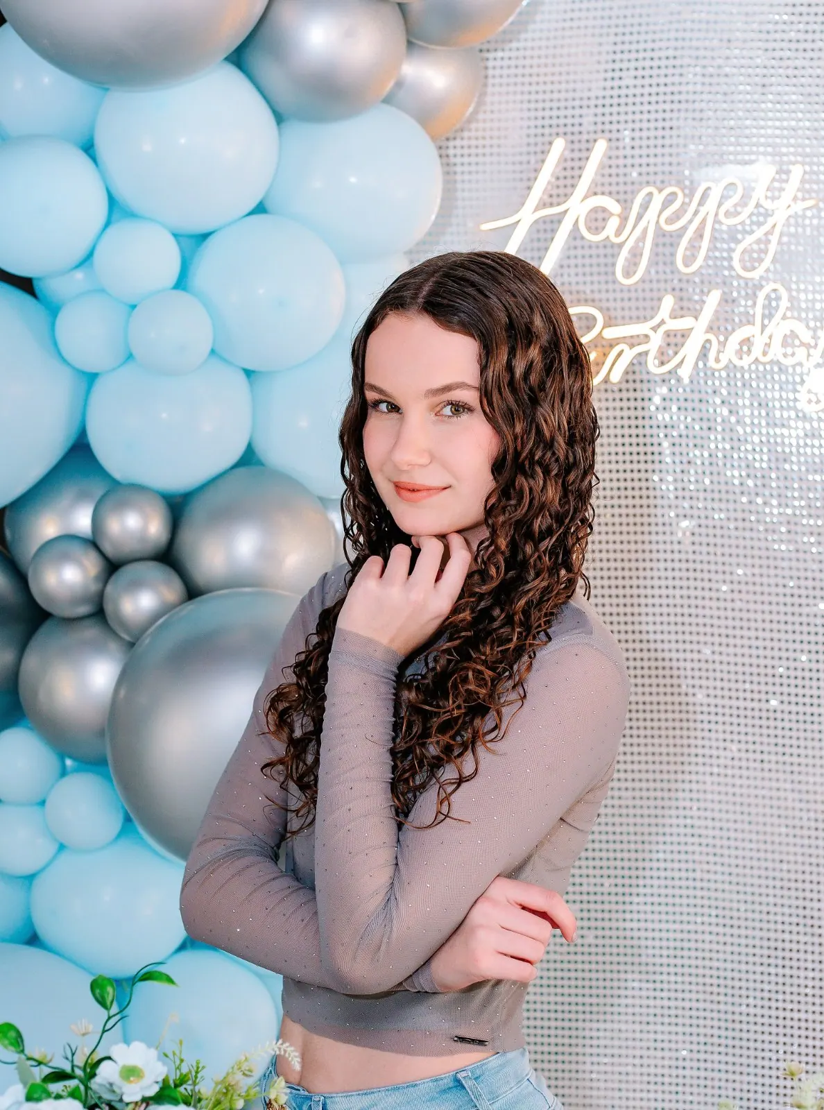 Adolescente de cabelos cacheados posa diante de balões azuis e letreiro Happy Birthday