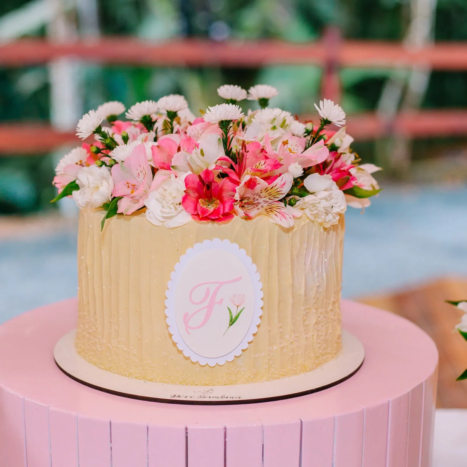 Bolo de aniversário coberto de flores naturais rosa e brancas sobre suporte rosa