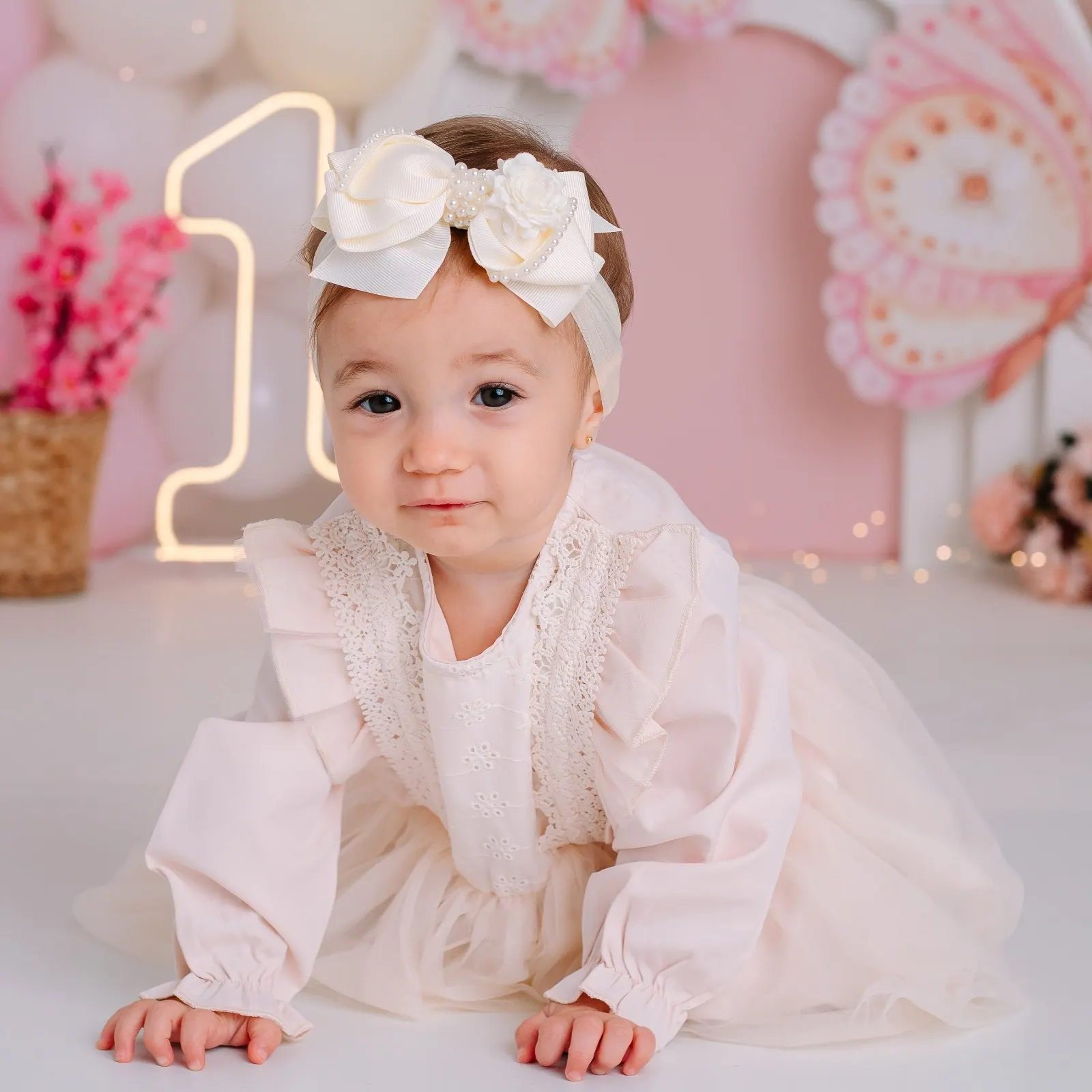 Bebê de laço creme e vestido de renda engatinha em cenário rosa de um ano