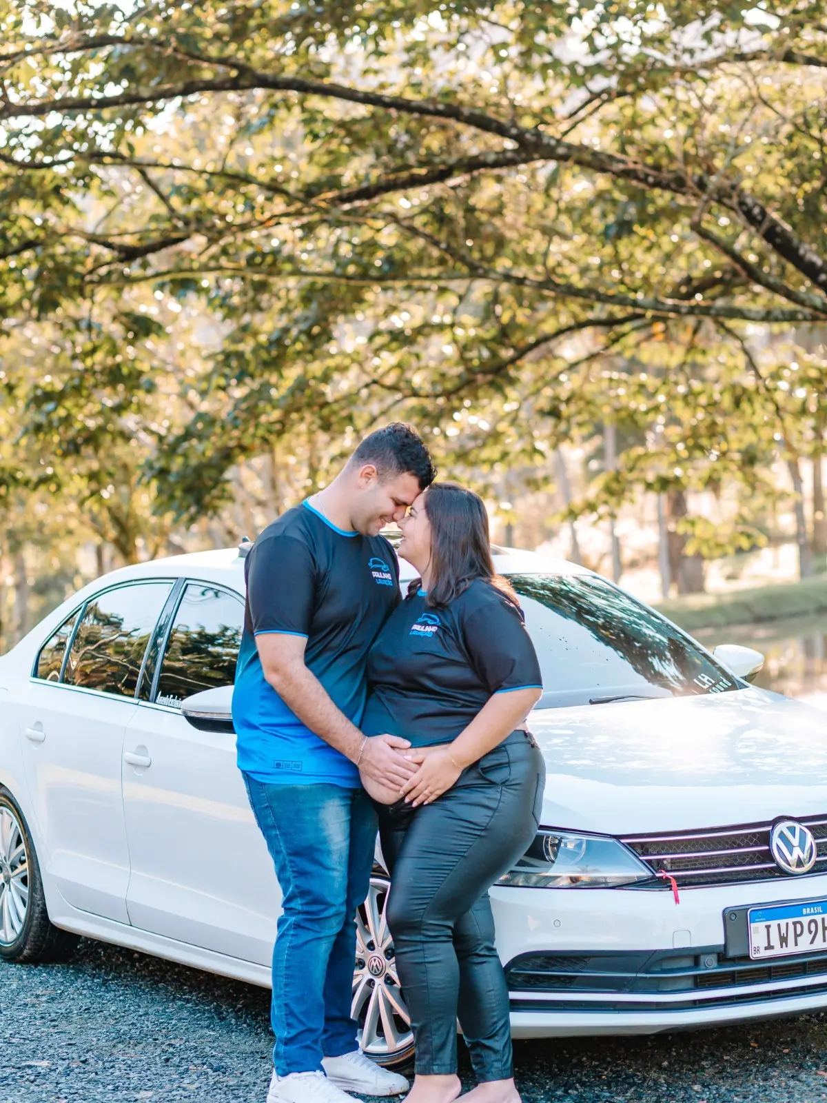 Casal encosta a testa um no outro com as mãos na barriga, sob árvores iluminadas pelo sol