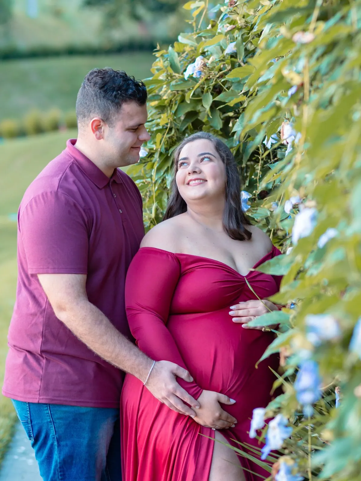 Casal em ensaio gestante ao ar livre, ela de vestido vinho sorrindo para ele entre flores