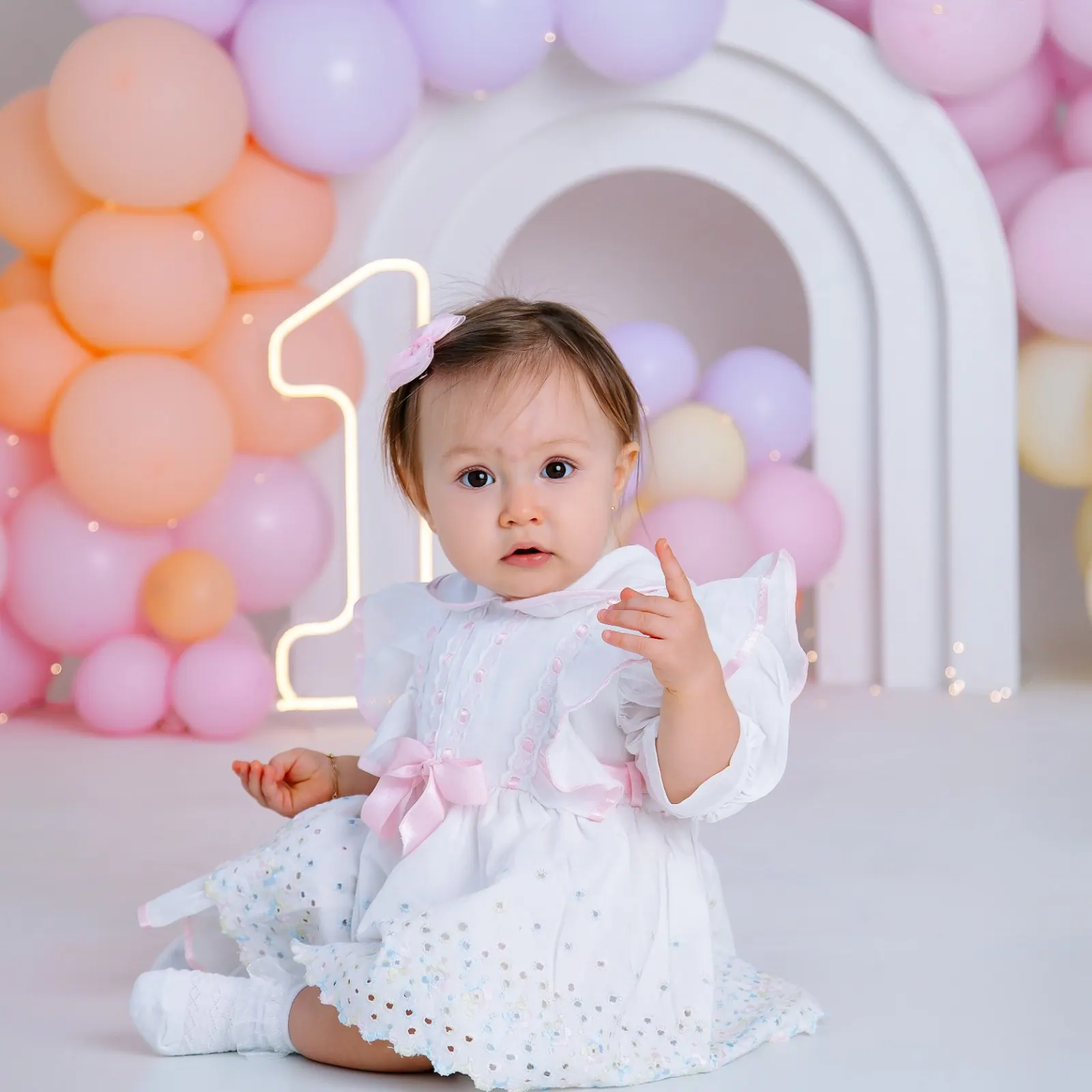 Bebê de vestido branco com laço rosa sentada diante de arco branco e balões em tons pastel