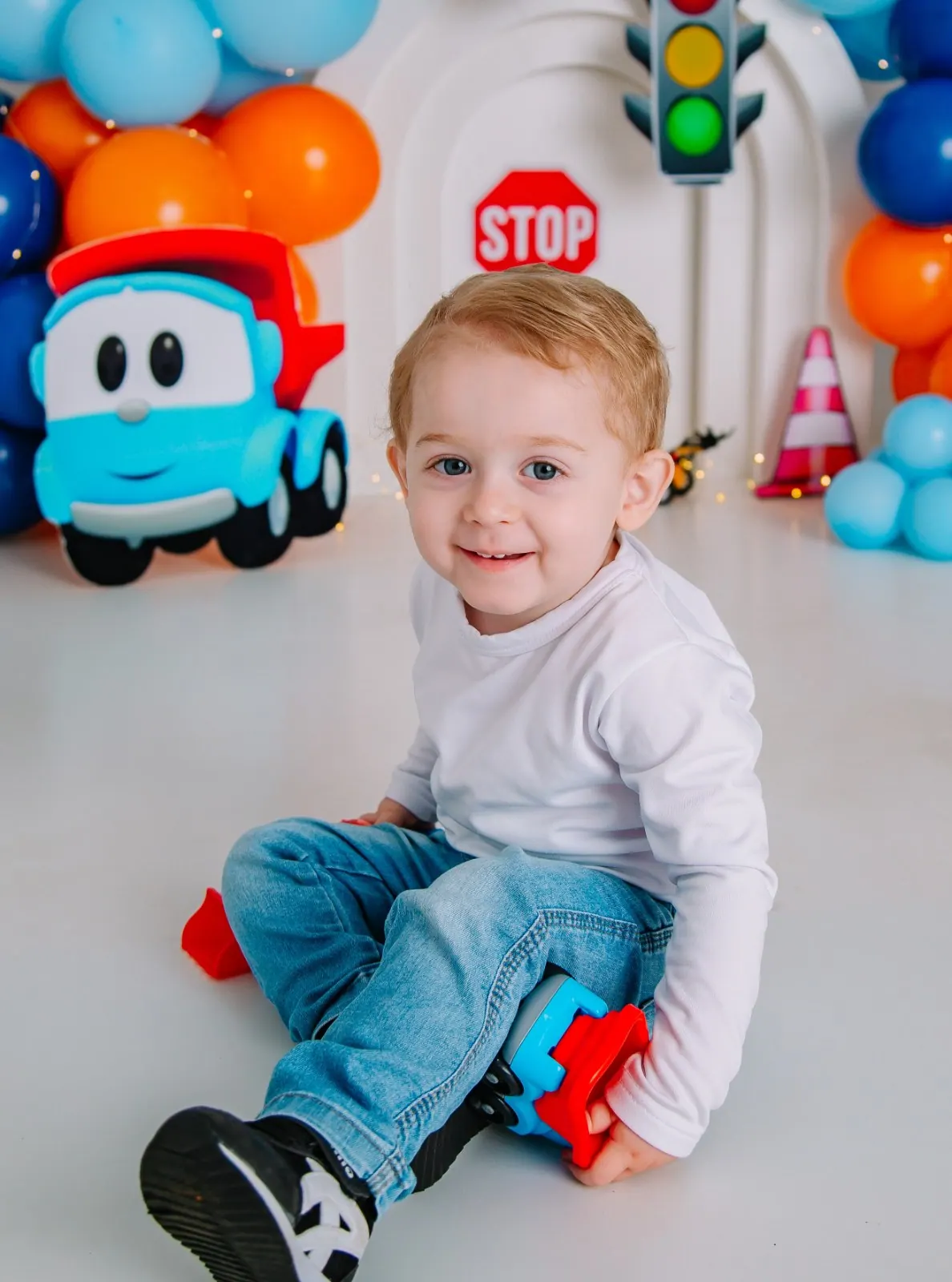 Menino sorridente sentado no chão com caminhão de brinquedo, balões azuis e laranja ao fundo