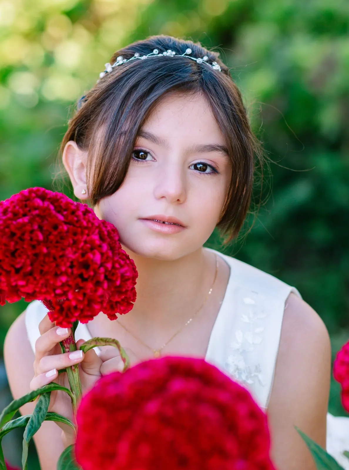 Menina com tiara de pérolas segura uma flor vermelha junto ao rosto, jardim desfocado ao fundo