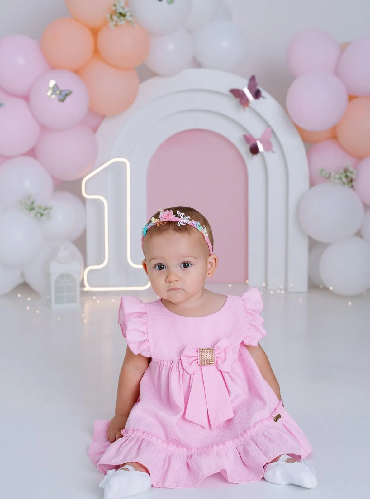 Bebê de vestido rosa com tiara de flores sentada diante de arco branco e número um iluminado