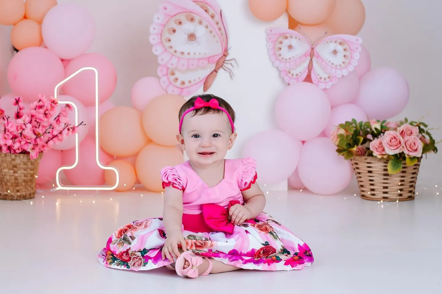 Bebê sorridente de vestido florido em cenário de borboletas e balões rosa